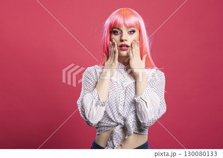 Attractive caucasian woman touching her cheeks and looking at camera with shocked facial expression. Surprised female individual standing with wide eyes and hands on face against pink background. 130080133