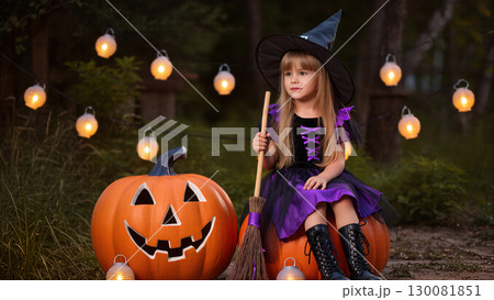 Little girl in witch costume sitting on pumpkin with broom during Halloween celebration with glowing lanterns. 130081851