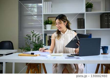 Young female accountant reviewing financial documents with clipboard and laptop in a home office Young female accountant reviewing financial documents with clipboard and laptop in a home office 130082460