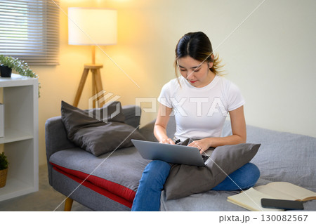 Young woman working remotely on laptop while sitting comfortably on sofa at home 130082567