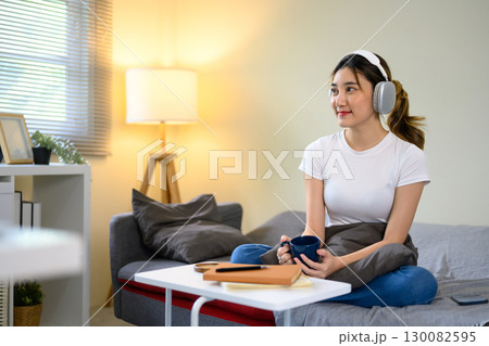 Relaxed young woman wearing headphones, sitting on sofa with coffee mug in a living room Relaxed young woman wearing headphones, sitting on sofa with coffee mug in a living room 130082595