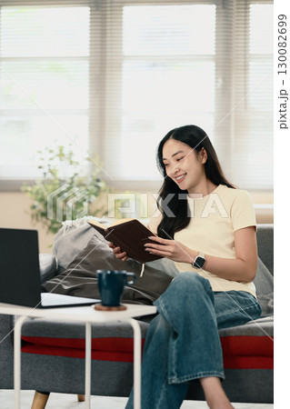 Smiling young woman sitting on sofa with notebook and working remotely at home Smiling young woman sitting on sofa with notebook and working remotely at home 130082699