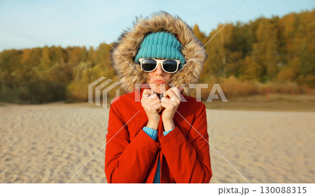 Woman is cold, freezing trying to warm up outdoors on beach on autumn day, wearing warm jacket Woman is cold, freezing trying to warm up outdoors on beach on autumn day, wearing warm jacket 130088315