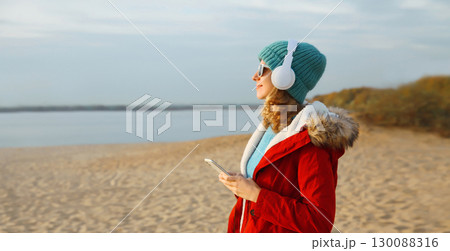Happy young woman enjoying listening to music in headphones with phone outdoors in autumn park 130088316
