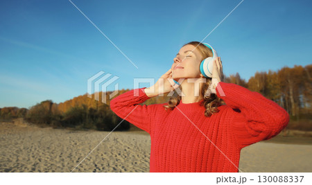 Happy young woman enjoying listening to music in headphones outdoors on autumn beach Happy young woman enjoying listening to music in headphones outdoors on autumn beach 130088337