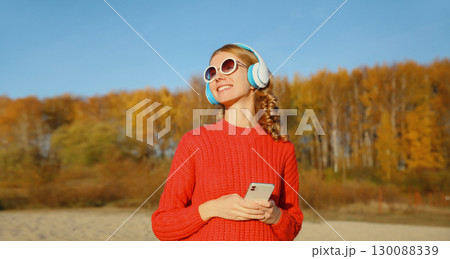 Happy young woman enjoying listening to music in headphones with phone outdoors in autumn park Happy young woman enjoying listening to music in headphones with phone outdoors in autumn park 130088339