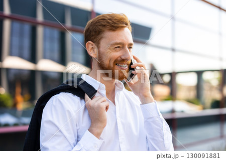 Red-haired man in white shirt standing outdoors talking on smartphone Red-haired man in white shirt standing outdoors talking on smartphone 130091881