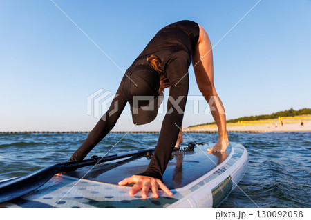 Young adult slim woman perform Yoga Downward-Facing Dog surfing SUP paddleboard Baltic Sea waves warm sunset sun lights summer day. Sunlight sea ocean waves enhance harmony body, strength mindfulness 130092058