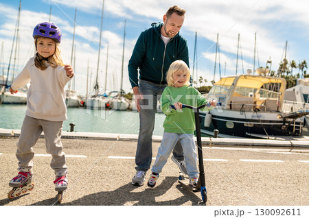 Parent supporting blond son riding scooter on dockside near yachts with skating daughter Parent supporting blond son riding scooter on dockside near yachts with skating daughter 130092611