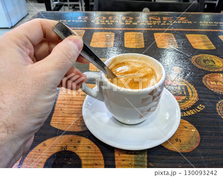 A person stirring a freshly brewed espresso in a white cup on a saucer, placed on a rustic wooden table. A person stirring a freshly brewed espresso in a white cup on a saucer, placed on a rustic wooden table. 130093242