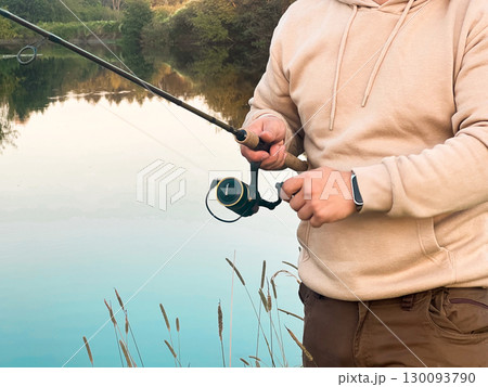 A young Caucasian man with short hair is fishing by a calm lake. He holds a fishing rod and reel, A young Caucasian man with short hair is fishing by a calm lake. He holds a fishing rod and reel, 130093790