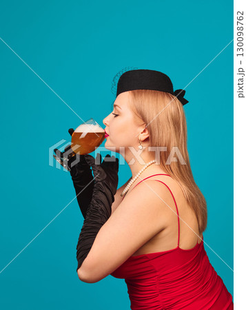 Woman in red dress with pearl necklace drinking beer glass in profile on blue background 130098762