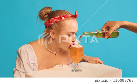 Woman drinking beer from glass while bottle pouring on blue background 130098765