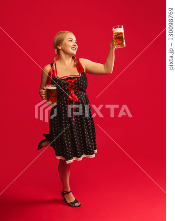 Happy woman in Oktoberfest dress raising two beer mugs with joyful smile against red background Happy woman in Oktoberfest dress raising two beer mugs with joyful smile against red background 130098769