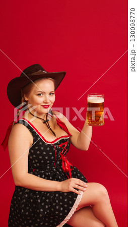 Woman in Bavarian dress with hat smiling and holding beer mug against red background Woman in Bavarian dress with hat smiling and holding beer mug against red background 130098770