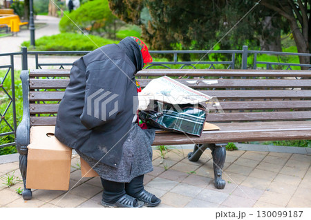 A homeless person in a red headscarf and black coat sits on a park bench, reading a newspaper A homeless person in a red headscarf and black coat sits on a park bench, reading a newspaper 130099187