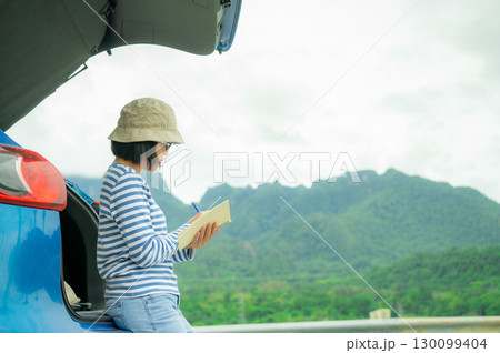 Woman writing in notebook for digital detox. Self-reflection, journaling habit, and mindful planning beside blue car with mountain landscape background. Road trip travel lifestyle. Mindful travel. 130099404