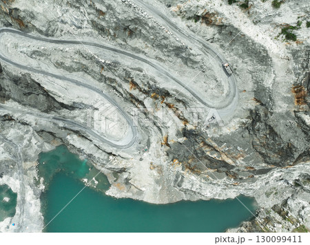 Aerial view truck in motion through terraced quarry levels during stone extraction. Mining technology, land use transformation, and corporate responsibility in heavy industry. Open pit mine industry. 130099411