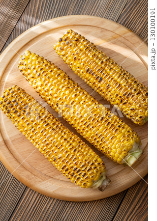 Three grilled corn cobs with char marks resting on wooden cutting board Three grilled corn cobs with char marks resting on wooden cutting board 130100191