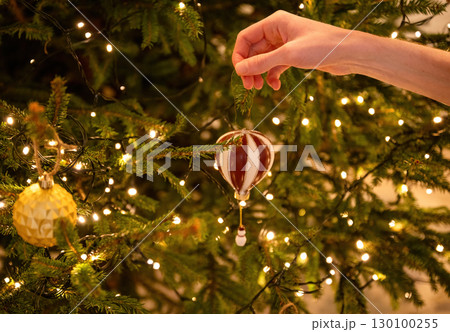 Hand hanging ornament on decorated Christmas tree with glowing lights 130100255