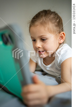Little girl lying on her parents bed and watching cartoons on a tablet. Concept of screen time, digital entertainment and relaxation during early childhood. 130101414