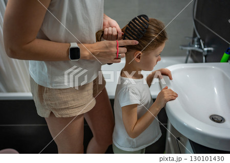 Mother braiding her daughter hair in the bathroom before going to school or kindergarten. Concept of morning preparation, care and family routine in childhood. Mother braiding her daughter hair in the bathroom before going to school or kindergarten. Concept of morning preparation, care and family routine in childhood. 130101430