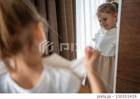 Little girl standing in front of a mirror and holding a dress against herself. Concept of self-expression, imagination and early interest in clothing and appearance. Little girl standing in front of a mirror and holding a dress against herself. Concept of self-expression, imagination and early interest in clothing and appearance. 130101439