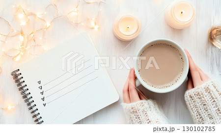 Cozy scene with woman holding warm drink, creating New Year goals resolutions list in notebook surrounded by candles and fairy lights 130102785