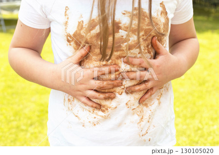Girl child staining her white t-shirt with stain while enjoying a delicious chocolate bar in garden Girl child staining her white t-shirt with stain while enjoying a delicious chocolate bar in garden 130105020