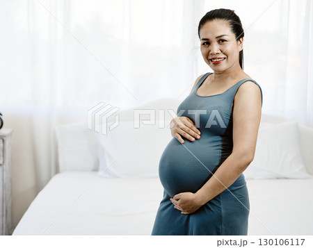 Pregnant woman in blue dress smiling while gently holding belly with both hands standing in bedroom with copy space 130106117