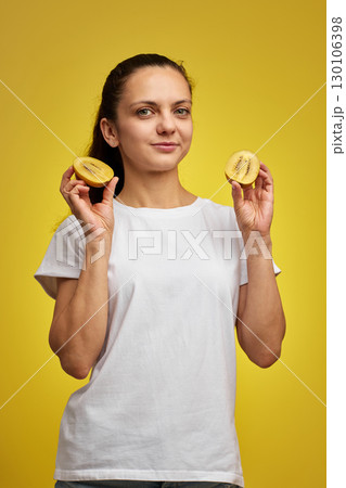 Happy woman in white t-shirt holding kiwi on studio yellow background 130106398