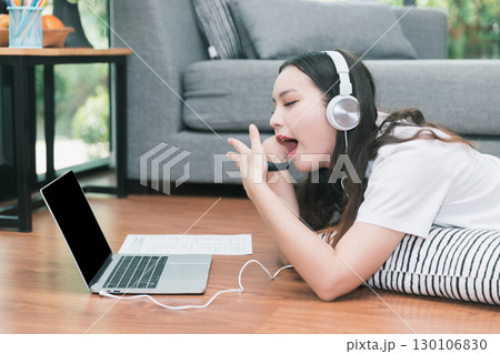 Young asian woman with headphone open mouth yawning while lying on the floor in living room. Studying online at home. Distance learning. Remote work place. Quarantine, coronovirus covid-19. 130106830