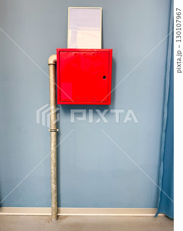 Red fire safety box on blue wall with metal pipe. Security, emergency, and urban infrastructure detail. Red fire safety box on blue wall with metal pipe. Security, emergency, and urban infrastructure detail. 130107967