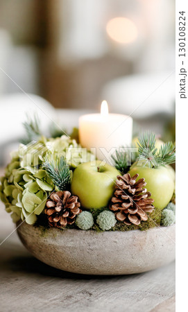 Decorative arrangement featuring green apples, pine cones, and hydrangeas in a rustic bowl with a lit candle creating a warm ambiance for home decor 130108224