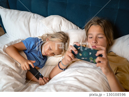 Two children lying in bed, using electronic devices for entertainment before sleep. Cozy setting with a focus on modern technology habits Two children lying in bed, using electronic devices for entertainment before sleep. Cozy setting with a focus on modern technology habits 130108596