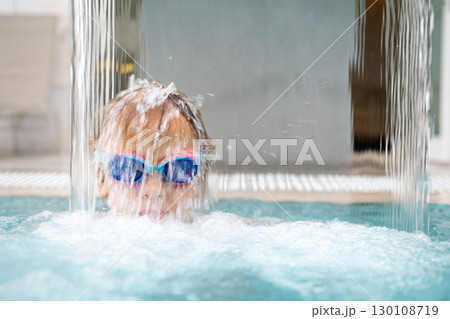 Blond child wearing swimming goggles enjoying refreshing waterfall in pool during summer vacation 130108719
