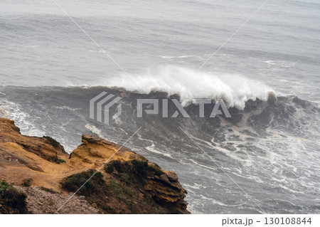 Massive ocean wave crashing, generating white foam near rocky cliff in Nazare, Portugal, renowned surfing destination with legendary swells Massive ocean wave crashing, generating white foam near rocky cliff in Nazare, Portugal, renowned surfing destination with legendary swells 130108844