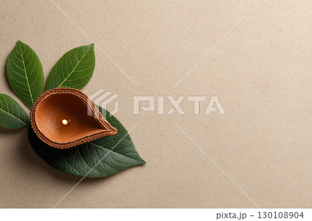 A small clay diya with a lit flame sits on green leaves on a textured beige surface. 130108904