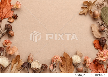 A flat lay of autumn leaves, acorns, and dried flowers arranged around the border of a light brown textured background. 130109023