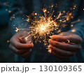 Woman holding a lit sparkler with both hands, creating a warm glow. 130109365