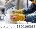 A person is putting the finishing touches on a small snowman wearing a hat in snowy weather. 130109368