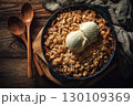A warm apple crumble topped with two scoops of vanilla ice cream in a round dish, placed on a wooden table with two wooden spoons and a cloth nearby. 130109369