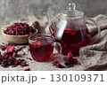 A glass teapot and cup filled with red hibiscus tea, surrounded by dried hibiscus petals on a textured fabric surface. 130109371