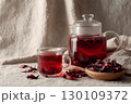 A glass teapot and cup filled with red hibiscus tea, surrounded by dried hibiscus petals on a textured fabric surface. 130109372