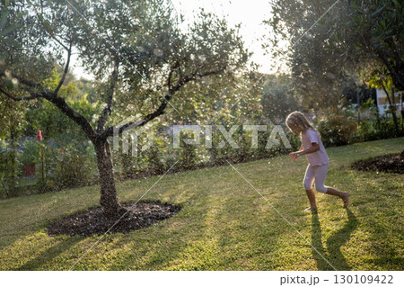 Playful kid splashing through sprinkler water, laughing amid verdant backyard greenery during sunny afternoon 130109422