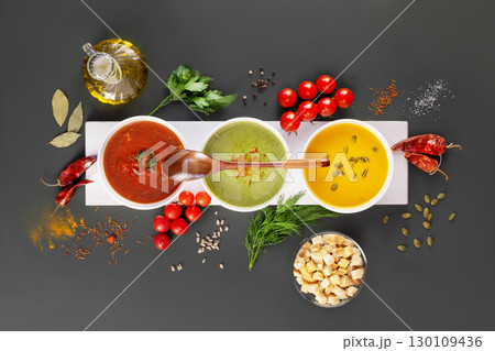 Set of different vegetable cream soups with wooden spoon on a white wooden board with ingredients on a dark grey background. Cozy meal, vegetarian healthy food. Top view 130109436
