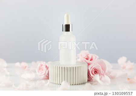 Glass dropper bottle with serum on a marble podium on a white table with tender pink roses and rose petals. Natural beauty product based on rose flowers, fermented cosmetic. Soft focus style 130109557