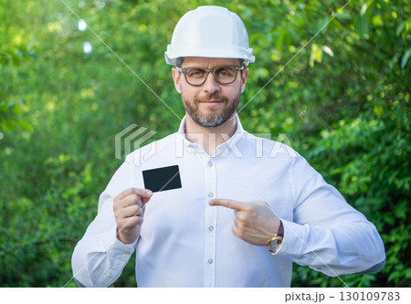 Businessman man in hardhat pointing finger at blank business card outdoors, copy space Businessman man in hardhat pointing finger at blank business card outdoors, copy space 130109783