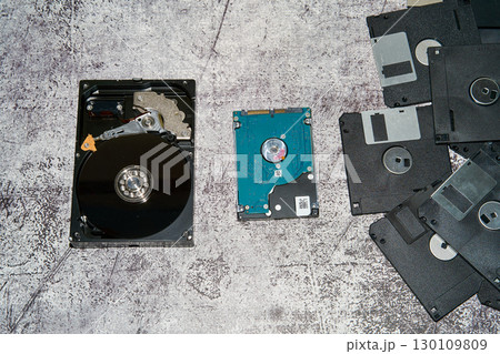 A 3.5 inch HDD with its casing open, a 2.5 inch HDD, and multiple floppy disks lie on a greyish backdrop A 3.5 inch HDD with its casing open, a 2.5 inch HDD, and multiple floppy disks lie on a greyish backdrop 130109809