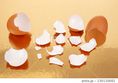 Two broken brown eggs with scattered eggshell fragments on a reflective golden surface 130110082